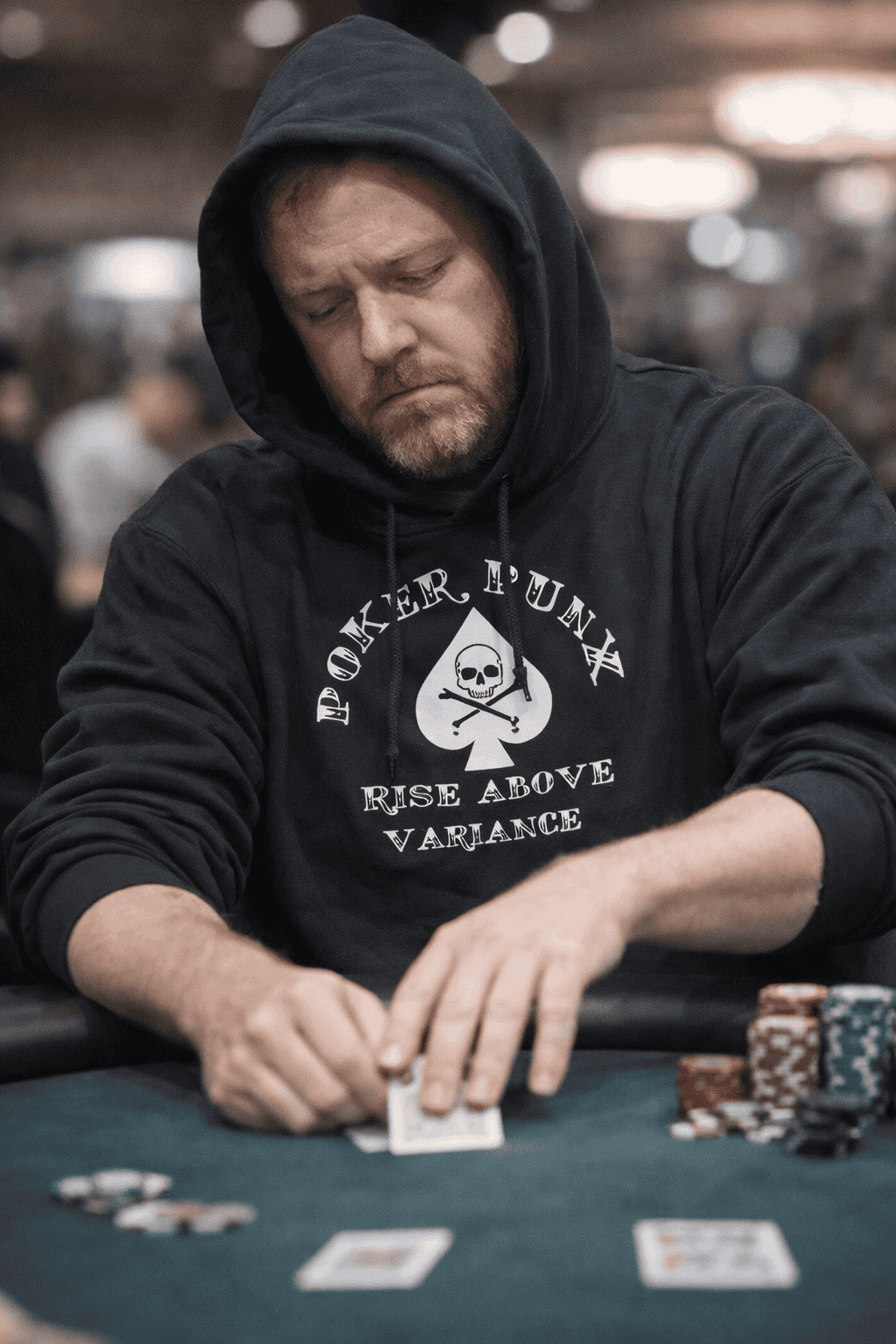 Poker player folding a hand with a resigned expression while wearing a Poker Punx Rise Above Variance hoodie at a casino table